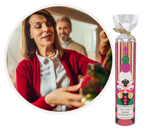 Frau in rotem Pullover dekoriert zuhause einen Weihnachtsbaum mit ihrem Partner im Hintergrund. Daneben eine bunte Weihnachtsschokoladenverpackung mit der Aufschrift 'Süßes Präsent Frohe Weihnacht'.