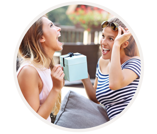 Zwei lachende Frauen, von denen eine ein Geschenk mit Schleife hält, draußen auf einer Terrasse sitzend