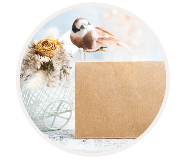 Ein dekoratives Arrangement aus einem künstlichen Vogel, Trockenblumen und einem leeren Karton auf einem Tisch.