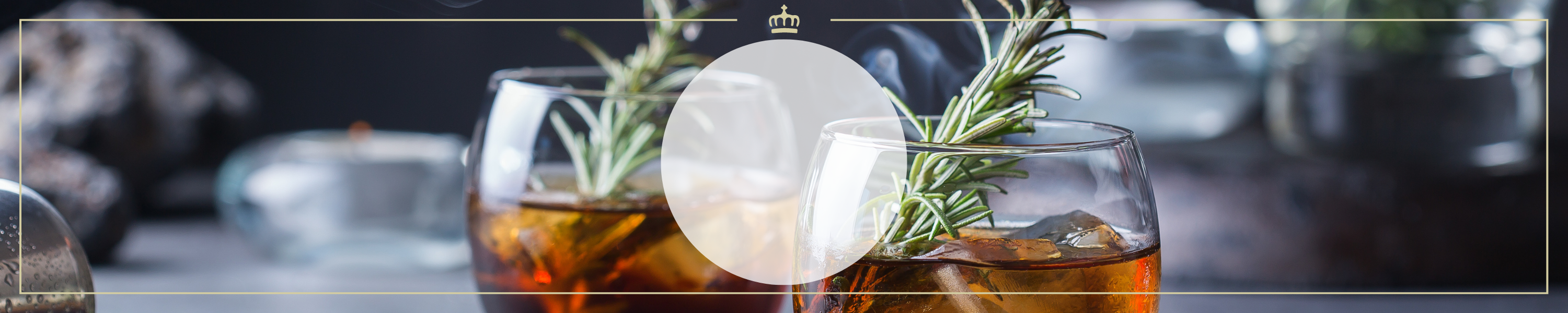 Ein Glas mit braunem Getränk, Eiswürfeln und Rosmarinzweig, von elegantem Ambiente umgeben