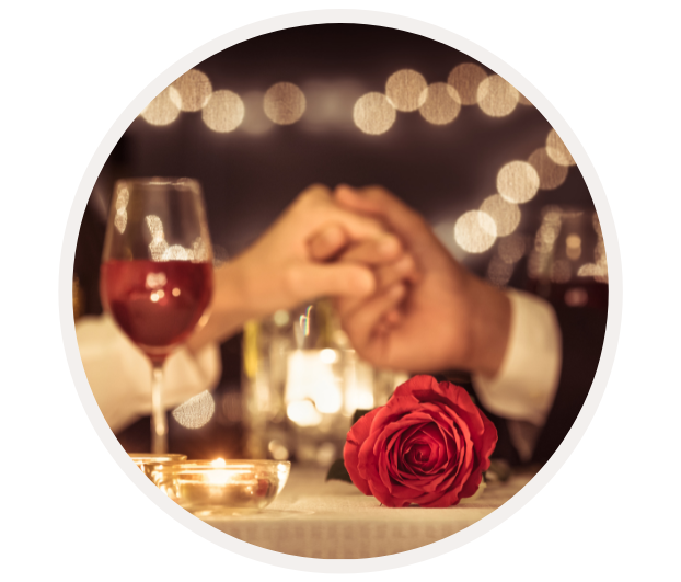 Paar hält sich bei einem romantischen Candle-Light-Dinner die Hände, umgeben von Rotwein, Kerzenlicht und einer roten Rose.