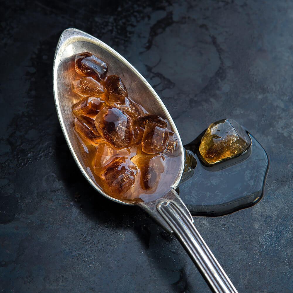 Rum-Kandis Braun in Großform auf Silberlöffel, braune Zuckerkristalle, daneben Vergleichsmuster auf dunklem Untergrund.