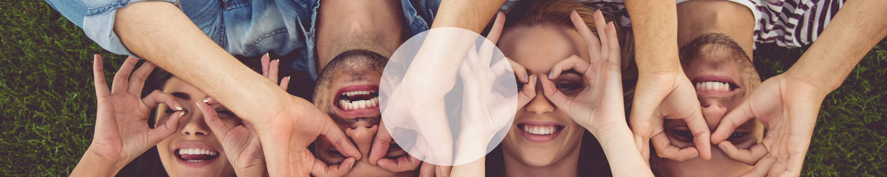 Lachende Freunde liegen auf dem Gras und formen mit ihren Händen Brillen vor ihren Augen.