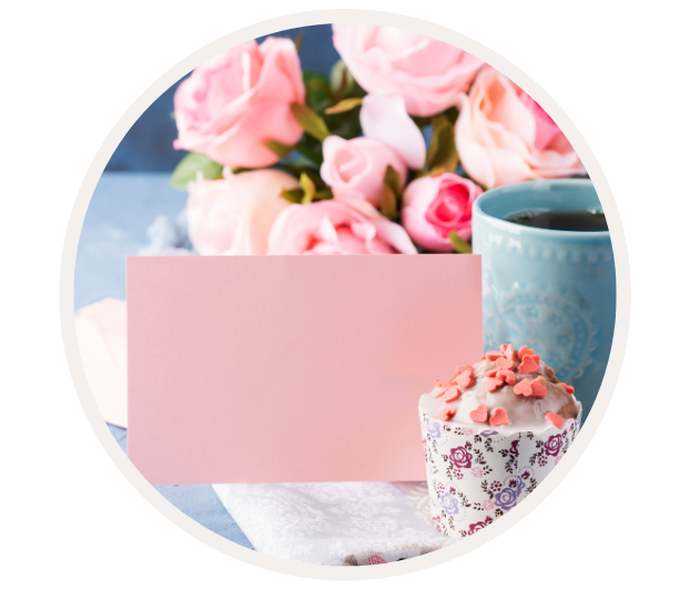 Ein Arrangement aus rosa Rosen, einer türkisen Tasse Kaffee, einem Cupcake mit Herz-Streuseln und einer leeren, rosa Karte auf einem Tisch.