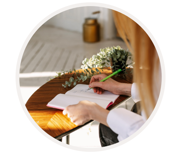 Eine Person schreibt mit einem grünen Stift in ein Notizbuch auf einem Holztisch neben einer Pflanze