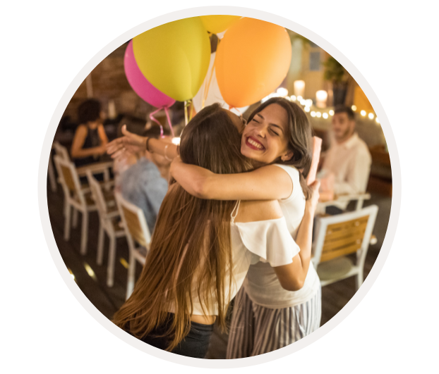 Zwei Frauen umarmen sich glücklich bei einer Feier, umgeben von Luftballons und festlicher Atmosphäre.