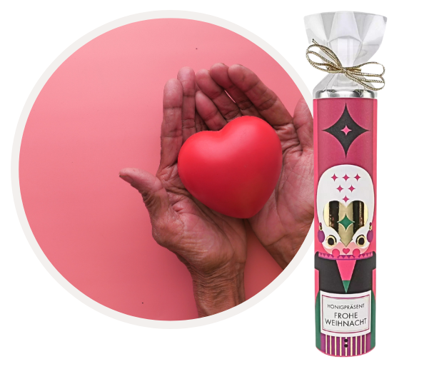 Eine Person hält ein rotes Herz in den Händen neben einer Verpackung mit der Aufschrift 'Honigpräsent Frohe Weihnacht'. Die Szene ist in einem rosa Hintergrund gehalten.