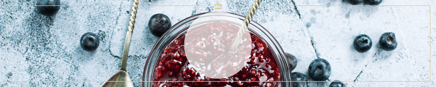 Ein Glas mit frischer Himbeermarmelade, umgeben von Heidelbeeren auf einem grauen Untergrund.