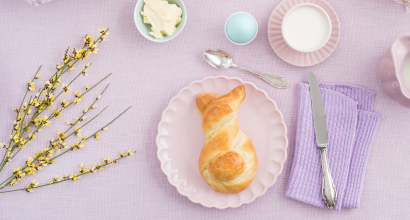 Eine stilvolle Tischdekoration mit einem katzenförmigen Gebäck auf einem rosa Teller, umgeben von Weidenkätzchen, Butter, einem Osterei, einer Tasse Milch und Löffel, Messer und Servietten auf lila Textilien.