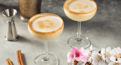 Zwei gläserne Cocktailgläser mit cremigen Getränken, Zimtstangen und Kirschblüten auf grauem Untergrund