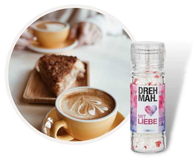 Nahaufnahme eines Cappuccinos mit herzförmigem Milchschaum, einer Scheibe Kuchen im Hintergrund und einer Packung DREHMAHL Zucker im Vordergrund