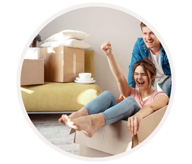 Lachende Frau sitzt in einem Umzugskarton, während ein Mann sie schiebt, umgeben von Kartons und Heimartikeln.