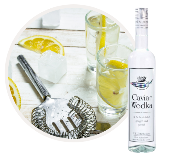 Flasche Caviar Wodka neben einem Cocktail mit Zitronenscheibe und Barzubehör auf weißem Holztisch