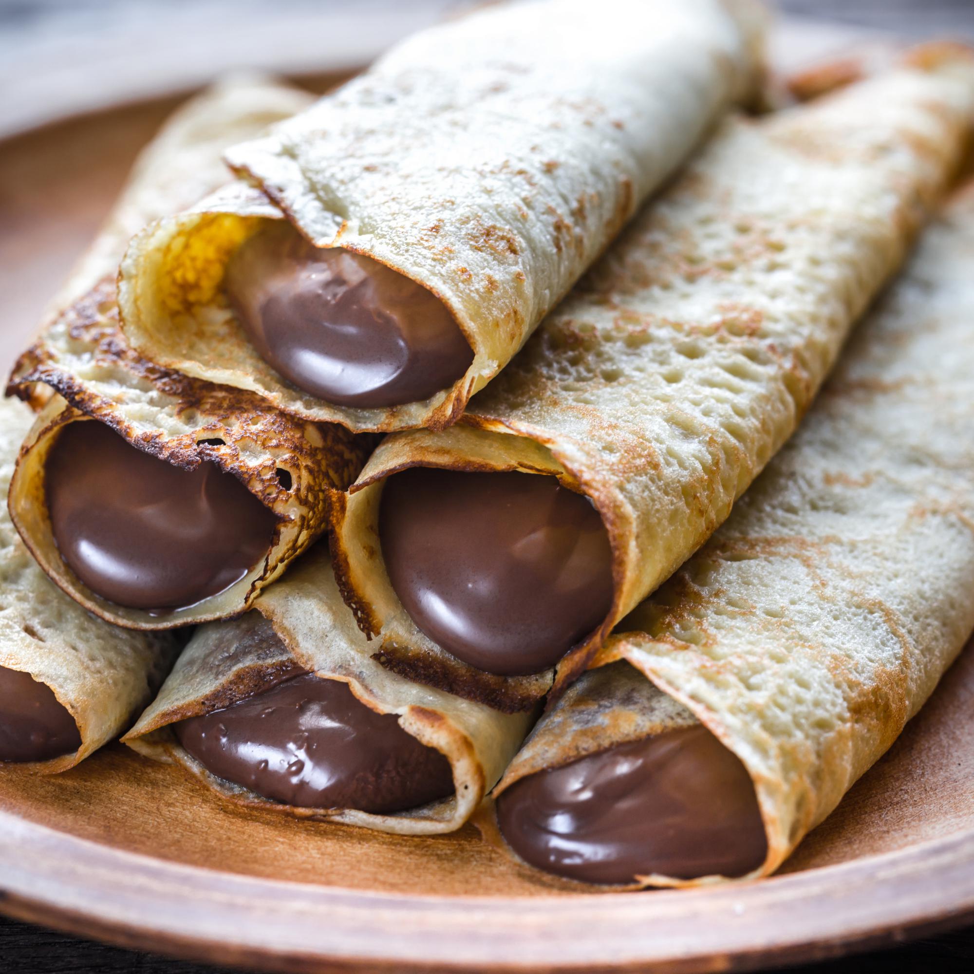 Nahaufnahme von mehreren gerollten Crêpes mit flüssiger Schokoladenfüllung auf einem Holzteller