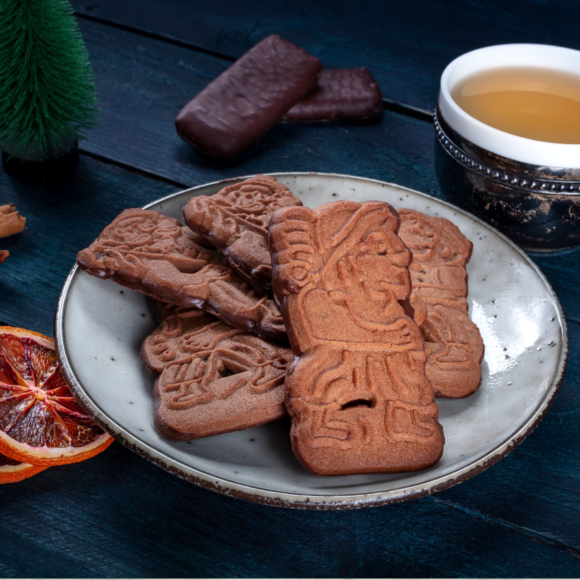 Ein Teller mit Spekulatius-Plätzchen neben einer Tasse Tee und Schokoladenkeksen, dekoriert mit getrockneten Orangenscheiben und einem kleinen Baum.