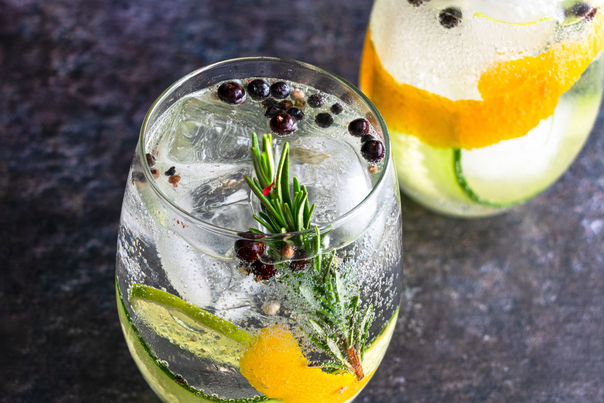 Ein Glas mit Gin Tonic, garniert mit Wacholderbeeren, Rosmarin und Zitrusfrüchten, neben einem weiteren Cocktailglas
