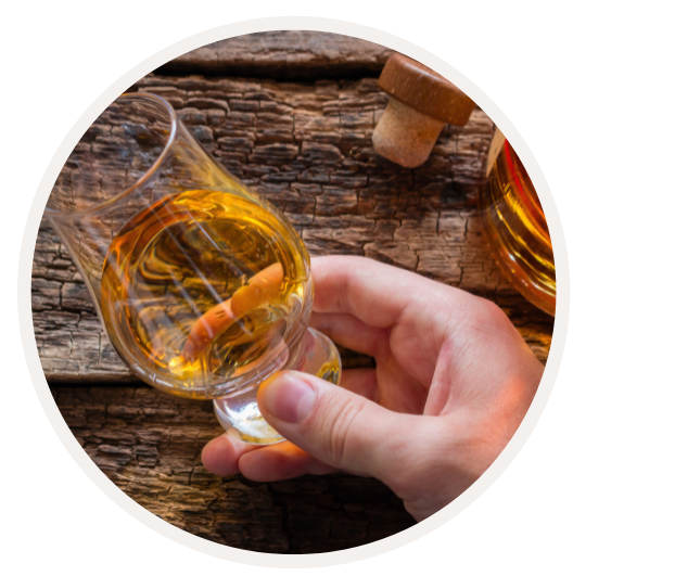 Nahaufnahme einer Hand, die ein Glencairn-Whiskyglas mit goldener Flüssigkeit auf einem Holztisch hält, daneben ein Korken.