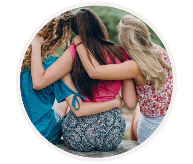 Rückansicht von drei Freundinnen, die einander umarmen und auf einer Bank im Freien sitzen. Sie tragen farbenfrohe Sommerkleidung.