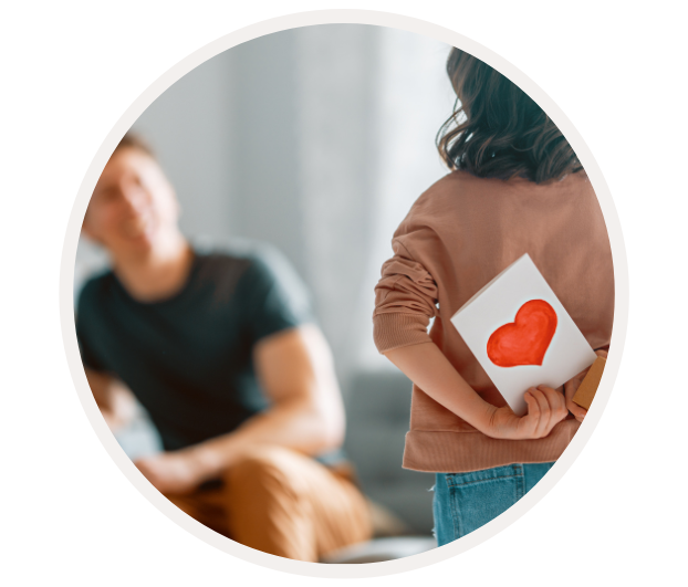 Ein Kind hält eine Karte mit einem roten Herz hinter dem Rücken, während ein Erwachsener im Hintergrund sitzt und zuschaut.