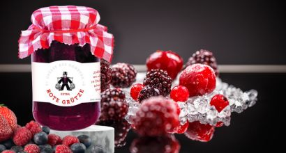 Ein Glas Rote Grütze mit einem rot-weiß karierten Deckel, umgeben von frischen Beeren und Eiswürfeln