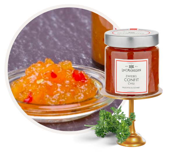 Ein Glas mit Zwiebelspezialität, fruchtig und scharf, neben einem Teller mit orangenem Confit und roten Chilistückchen, verziert mit frischem Kraut.