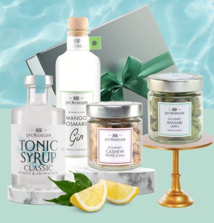 Ein Geschenkset bestehend aus Mango Rosmarin Gin, Tonic Sirup, Cashewkernen mit Trüffel und Salz sowie Wasabi Erdnüssen, arrangiert vor einem eleganten Hintergrund.